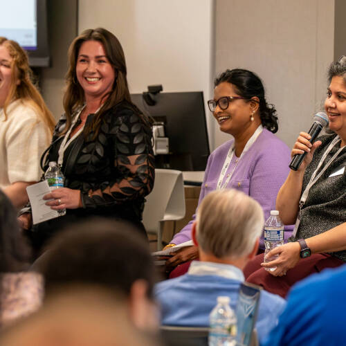 four women sit on tall chairs, person on right with microphone during a panel discussion; audience seated in front